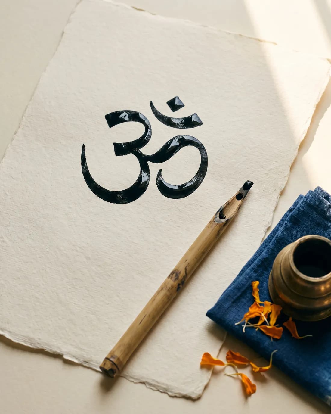 The Devanagari ॐ freshly written in black ink on cream handmade paper, a cut bamboo reed pen beside it, a brass inkwell and marigold petals at the edge