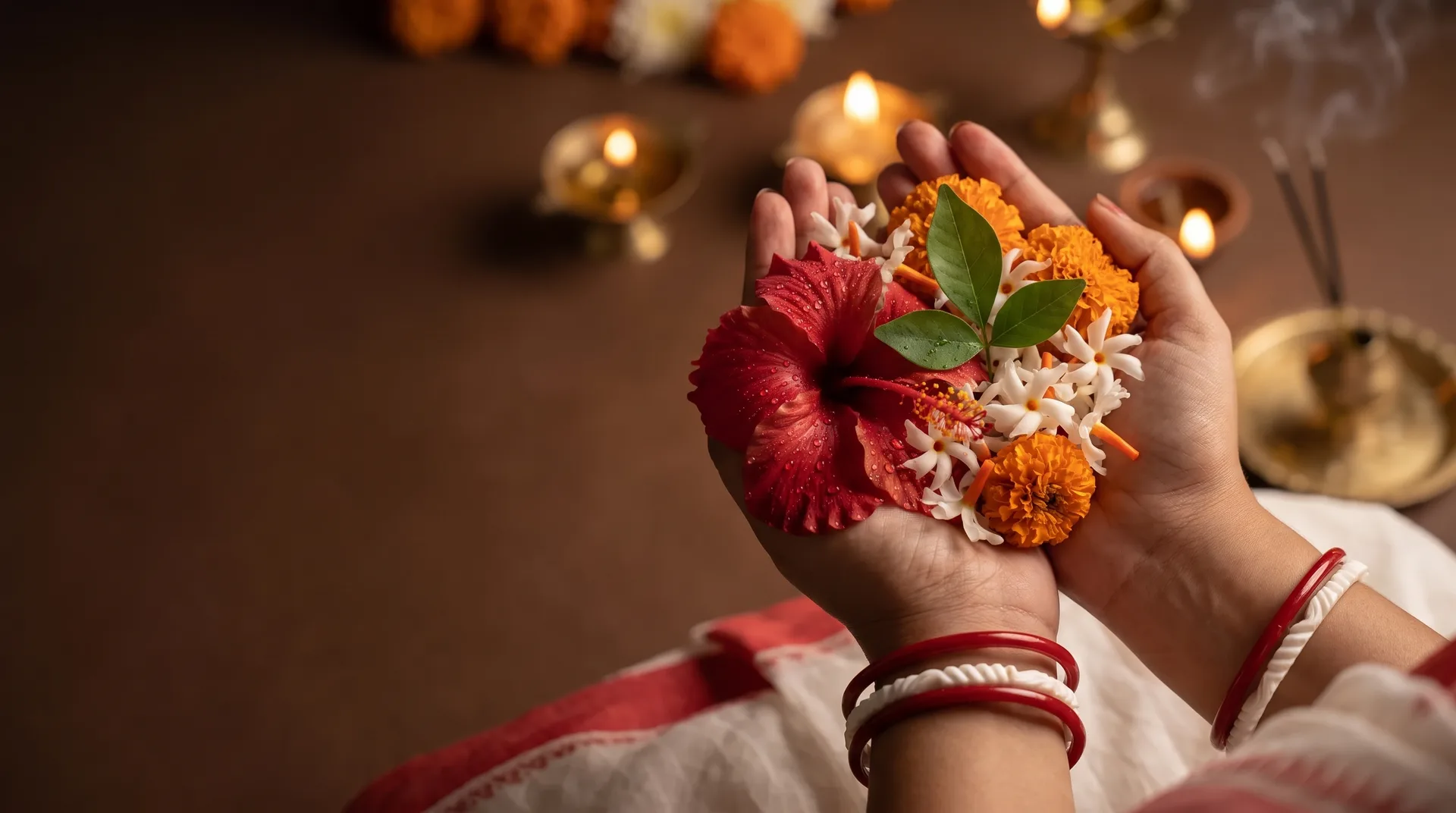 Two cupped hands holding a pushpanjali of red hibiscus, white shiuli with orange stems, marigold and a bilva leaf, at a Durga Puja morning